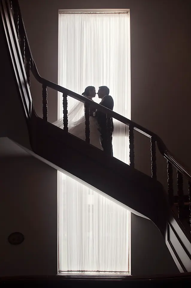 Silueta romántica de los novios a punto de besarse en una escalera oscura, enmarcados por la luz blanca de un gran ventanal al fondo. Wilder Fotografia